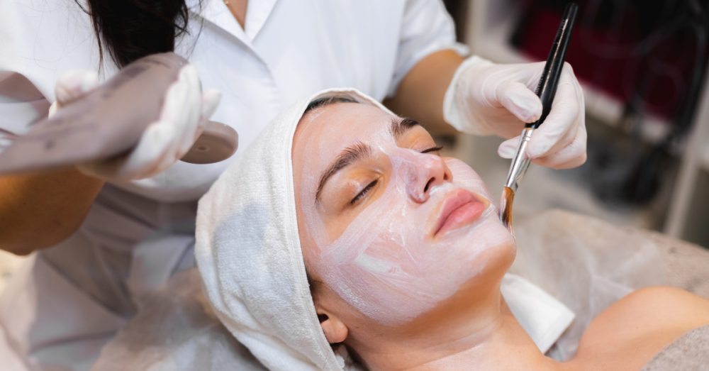 A gloved hand applying a white facial mask with a brush to a woman lying down wearing a head wrap