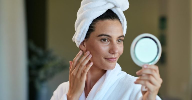 A woman wrapped in a white towel and robe holding a small mirror while touching her cheek and smiling